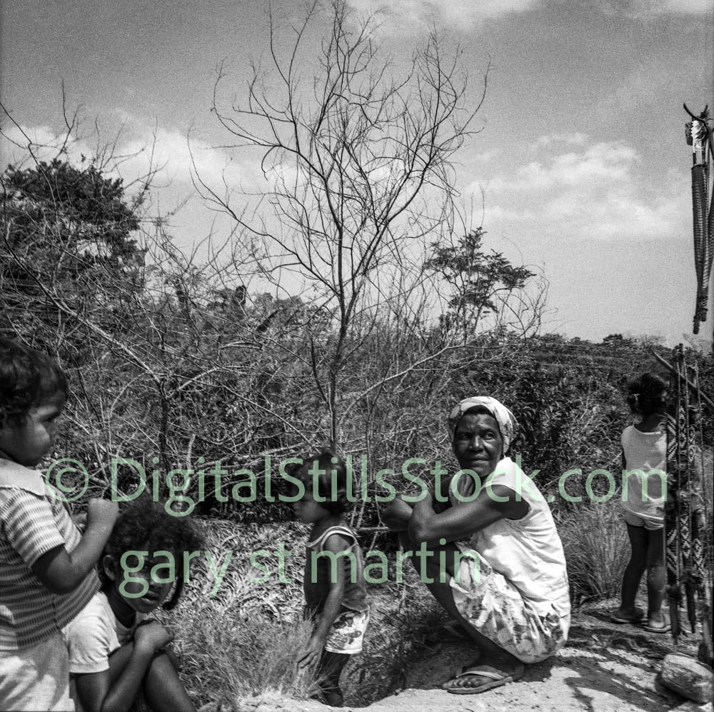 Black and white photograph of a woman and children outdoors with trees and sky in the background.