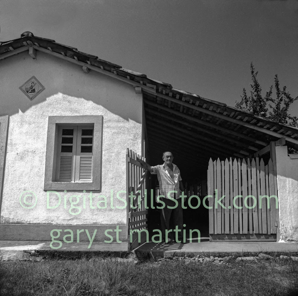Black and white photo of a man standing in front of a small building with a picket fence.