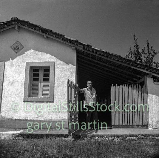 Black and white photo of a man standing in front of a small building with a picket fence.