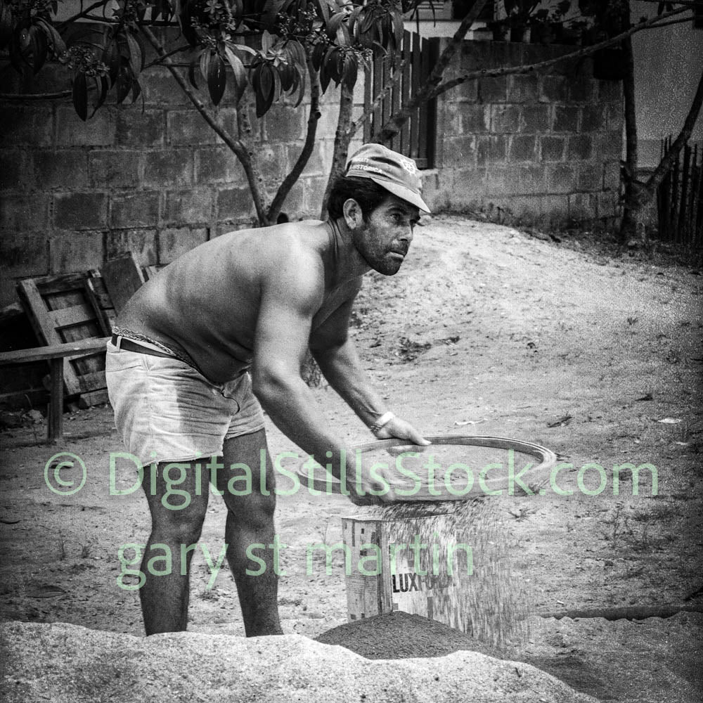 Man in a hat and shorts working with sand or gravel in an outdoor setting.