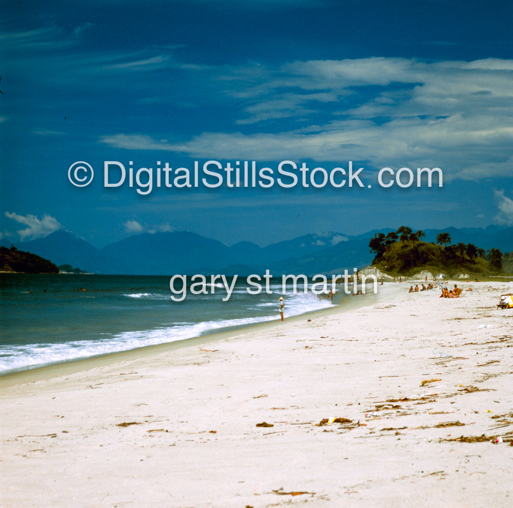 Beach scene with ocean, sand, and mountains under a blue sky.